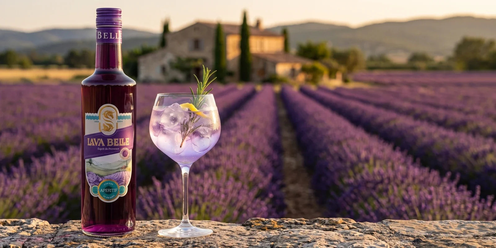 Flasche Lav'a Belle Lavendel-Aperitif auf einer Steinmauer vor einem Lavendelfeld in der Provence, daneben ein Spritz-Glas mit Eis und Rosmarin